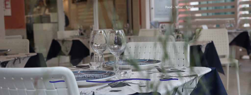 Table set with plates and glasses in a restaurant setting, featuring white chairs and patterned tablecloth.