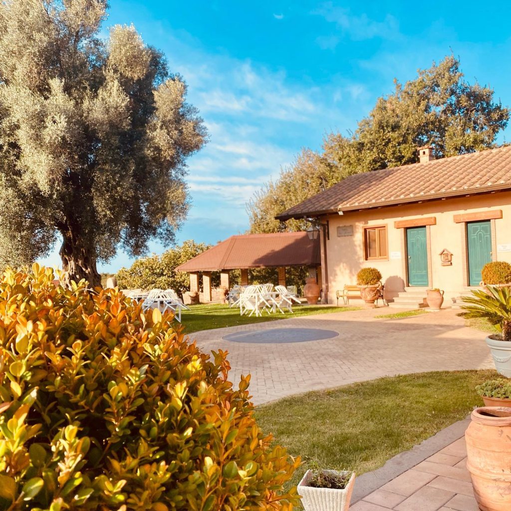 A rural Italian house with a garden featuring green grass and ornamental plants under a blue sky.