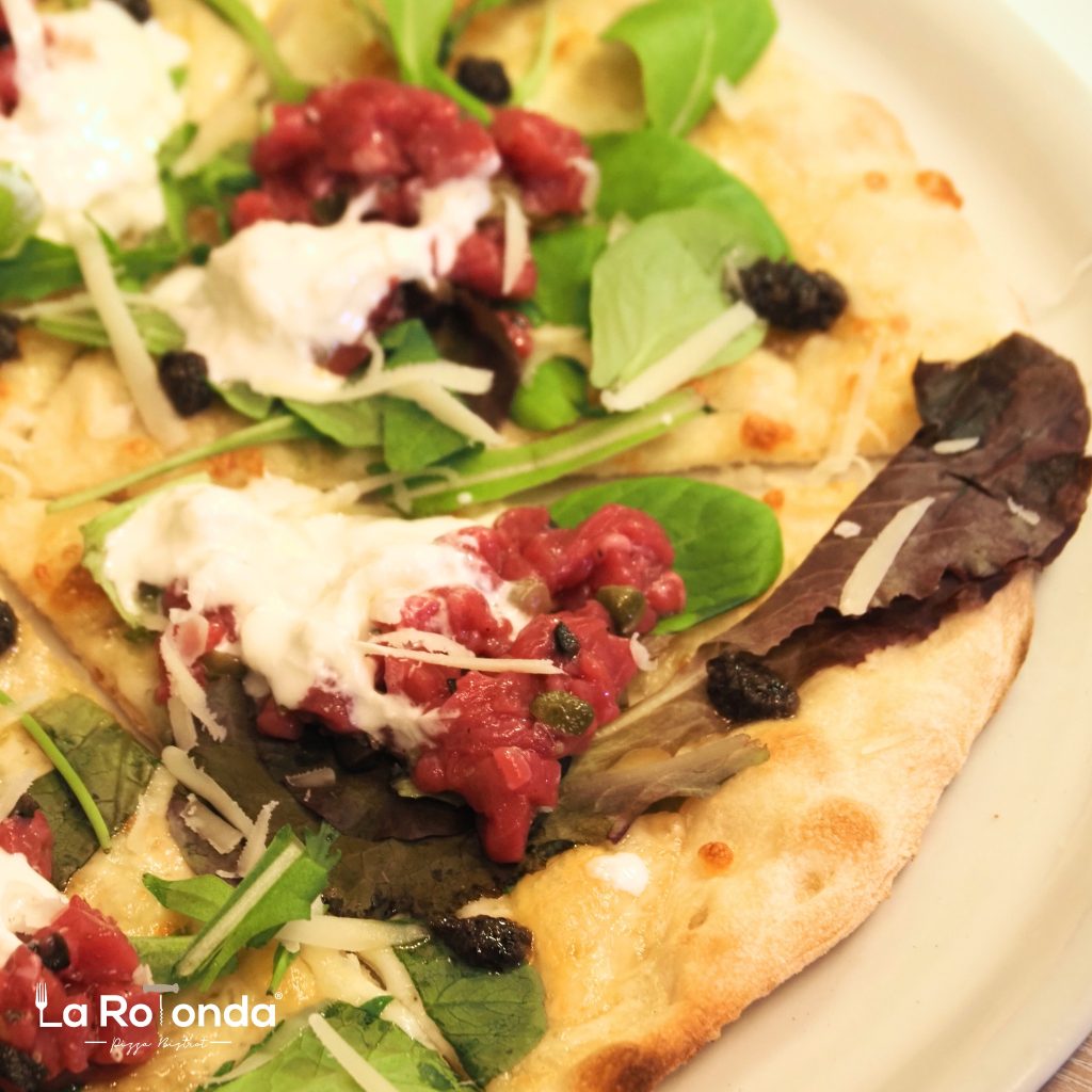 Pizza topped with beef tartare, greens, and dollops of cream on a crust.